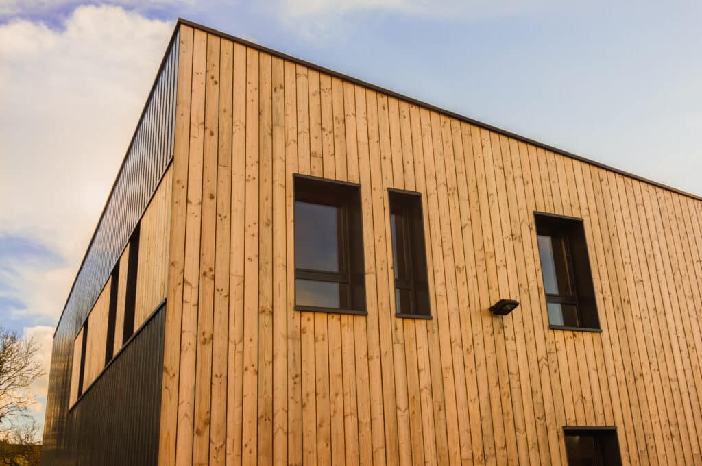 Façade angulaire d'un bâtiment moderne : bois clair vertical, bardage métallique noir. Fenêtres sombres, éclairage sous ciel nuageux.