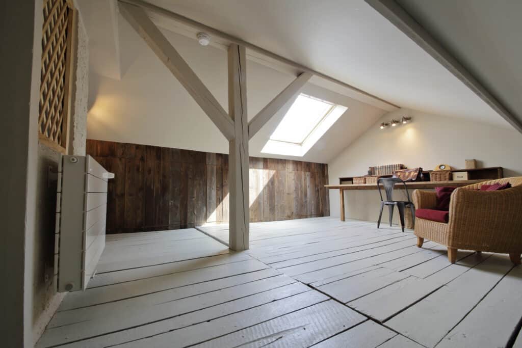 Bureau lumineux sous les combles avec poutres apparentes, plancher bois clair, mur en bois foncé, et lucarne. Fauteuil en osier.