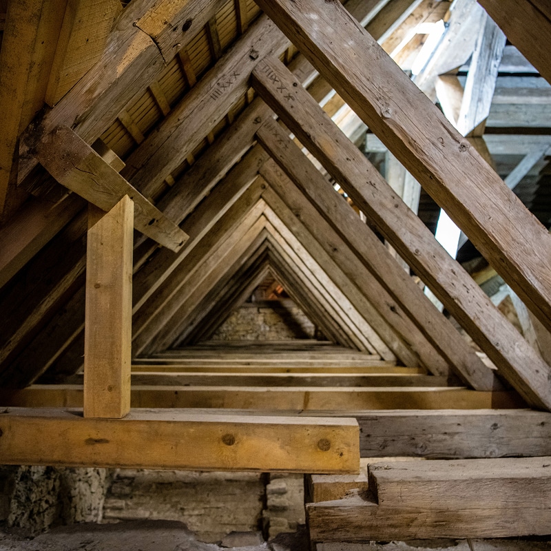 Charpente ancienne en bois brut d'un grenier, créant une perspective triangulaire profonde. Marques de charpentier visibles sur les poutres.