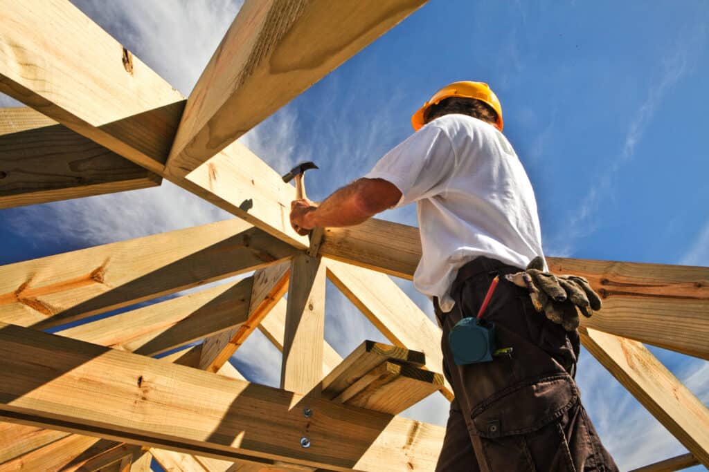 Charpentier en plein travail sur une charpente en bois de toit, tenant un marteau, sous un ciel bleu ensoleillé.