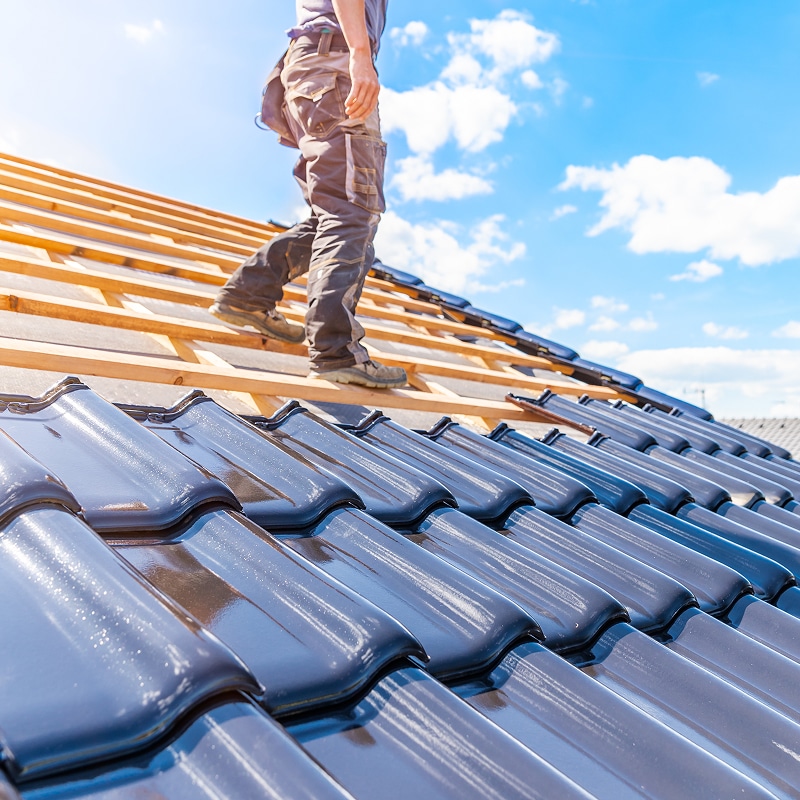 Couvreur marchant sur la charpente en bois d'un toit en construction. Des tuiles noires brillantes sont posées au premier plan.