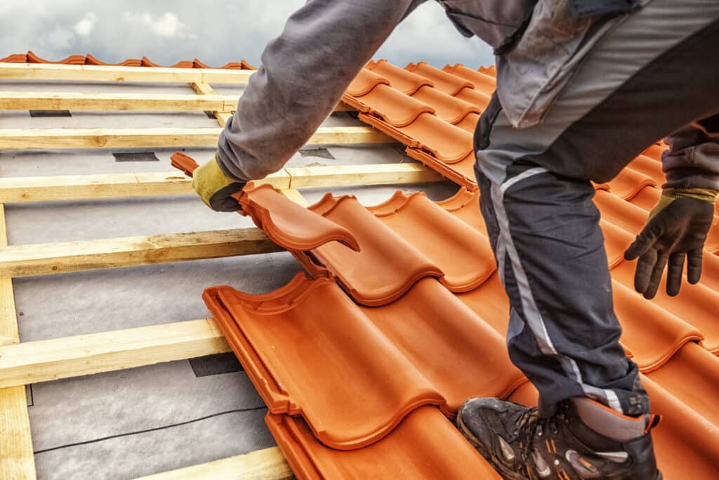 Couvreur pose des tuiles en terre cuite sur une toiture en construction, avec des liteaux en bois et une bâche grise. Travaux de couverture.
