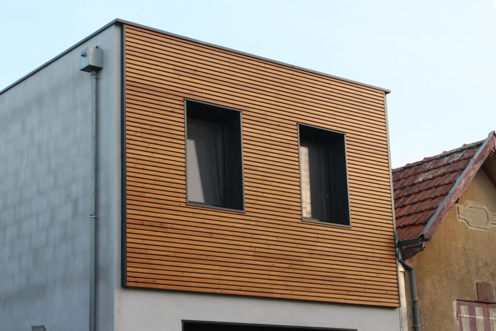 Façade supérieure d'une maison moderne avec bardage en bois clair et deux fenêtres noires, juxtaposée à un toit de tuiles rouges d'une bâtisse ancienne.