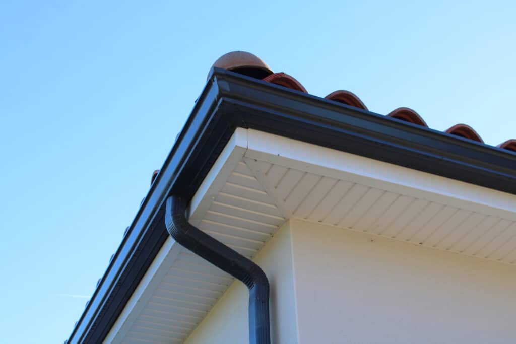 Coin de maison avec gouttière noire, tuiles de toit en terre cuite et soffite blanc sous un ciel bleu clair.
