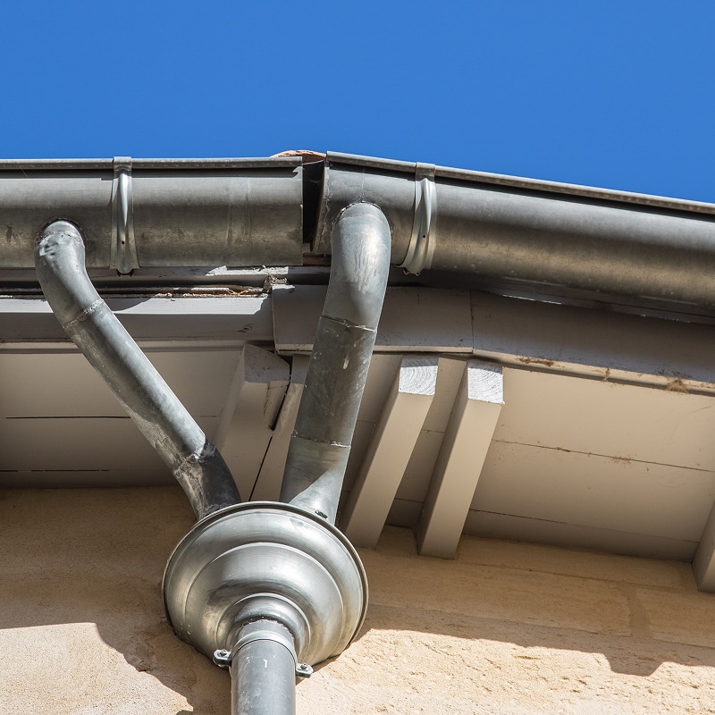Gouttière et descente d'eau en zinc sur la façade d'un bâtiment ancien sous ciel bleu. Deux tuyaux convergent vers une tête de moine.