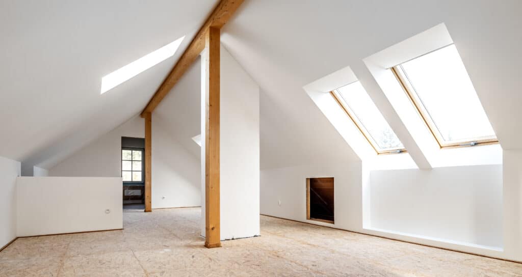 Grand loft mansardé lumineux en cours de rénovation avec murs blancs, poutres en bois, lucarnes et plancher OSB.