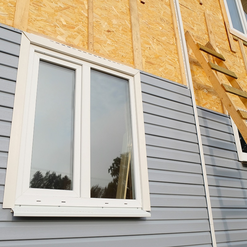 Façade de maison en construction avec bardage vinyle gris, fenêtre blanche installée et structure en panneaux OSB apparente.
