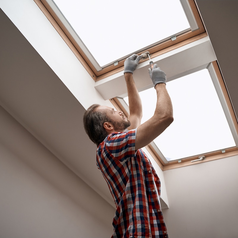 Homme en chemise à carreaux et gants répare une lucarne avec un tournevis. Entretien de fenêtre de toit dans une maison lumineuse.