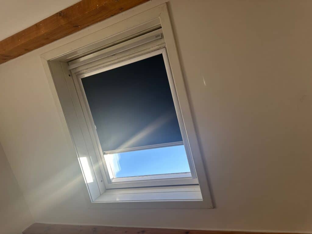 Fenêtre de toit Velux: Lumière naturelle et store occultant Fenêtre de toit Velux avec store noir semi-ouvert, laissant entrer soleil et ciel bleu. Rayons lumineux sur mur beige, poutres bois apparentes.